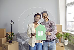 Happy young black couple standing in their new home, holding paper house and smiling