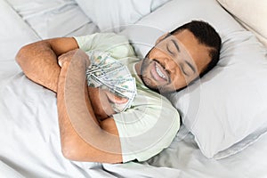 Happy young black guy hugging money cash, laying in bed