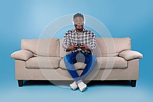 Happy young black guy with joystick playing video game, wearing headset, sitting on couch over blue studio background