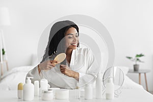 Happy young black lady in bathrobe sitting in bedroom, combing her curly hair with wooden brush, copy space