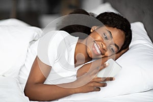 Happy young black lady laying in bed with smartphone