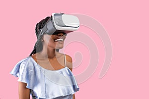Happy young black lady wearing VR headset, exploring cyberspace, using virtual reality on pink background, copy space