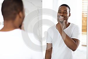 Happy Young Black Man Looking In Mirror In Bathroom And Touching Face