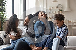 Happy young Black parents cuddling, tickling kids on couch