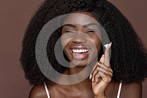 Happy young black woman applying facial cream isolated on brown background.