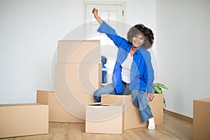 Happy Young Black Woman Celebrating Moving Home, Sitting On Big Cardboard Box