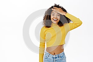 Happy young black woman, excited girl holds forehead, looks surprised and excited, receive great news, stands over white