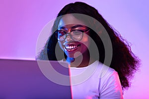 Happy young black woman in glasses using laptop pc for online work or studies in neon light