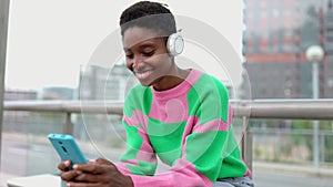 Happy young black woman with headphones using cell phone sitting on tram station