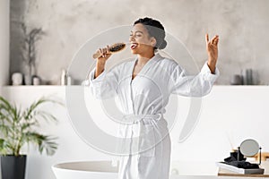 Happy young black woman holding hairbrush like microphone while singing in bathroom
