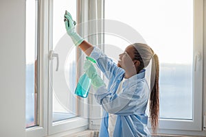 Happy young black woman housekeeper cleaning windows at apartment