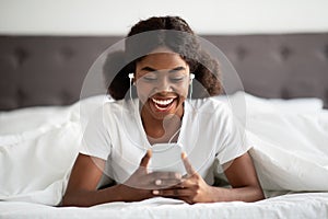 Happy young black woman laying in bed with smartphone, earphones