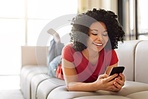 Happy young black woman messaging on smartphone while relaxing at home