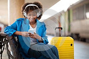 Happy Young Black Woman Messaging On Smartphone While Sitting At Railway Station