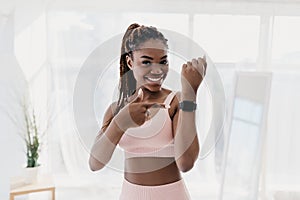 Happy young black woman pointing at activity fitness tracker with mockup, having break in her training at home