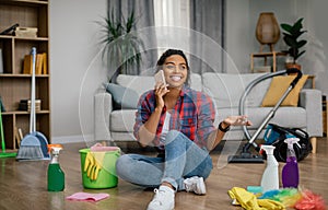 Happy young black woman in rubber gloves sit on floor with cleaning supplies, calling by phone in living room