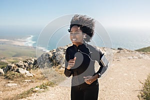 Happy young black woman running in morning, training on rocks with ocean view, enjoying workout outdoors