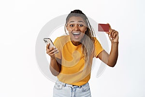 Happy young black woman showing her credit card after using mobile phone app, laughing with joyful face expression