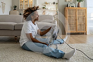 Happy young black woman sits in front of fan at home cooling down at home after being in heat