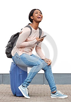 Happy young black woman sitting on suitcase and laughing with mobile phone