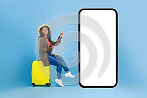 Happy young black woman sitting on suitcase and pointing at smartphone with mockup for online travel agency website