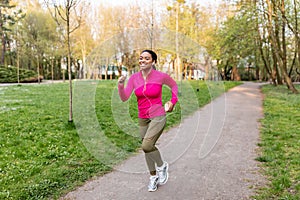 Happy young black woman in sports outfit running with mobile device and earphones at park, full length