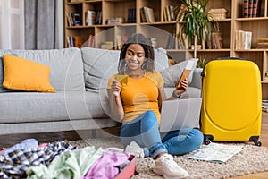 Happy young black woman using credit card and laptop, booking abroad trip, holding passport with tickets