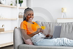 Happy young black woman using laptop and credit card, checking new website for online shopping or banking at home