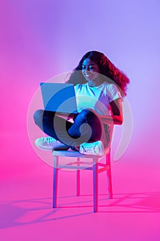 Happy young black woman using laptop pc for online work or studies, sitting on chair in neon light, full length