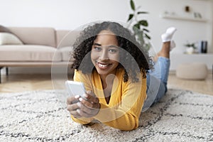 Happy young black woman using mobile phone, having online call, lying on floor, posting in social media at home