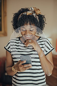 Happy young black woman using a smartphone indoors wearing wireless earbuds