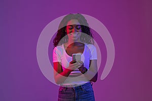 Happy Young Black Woman Using Smartphone While Standing In Bright Neon Light