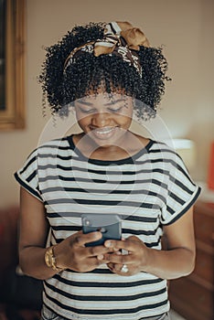 Happy young black woman using a smartphone wearing wireless earbuds at home