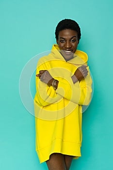 Happy young black woman in vibrant yellow oversized hooded sweatshirt is posing with arms crossed