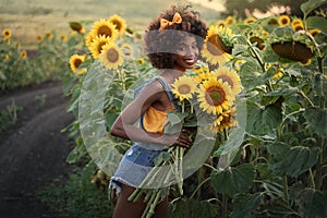 Happy young black woman walks in the sunflower field. Smiling dark-skinned girl with with a bouquet of sunflowers and