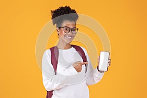 Happy young black woman wearing glasses and backpack pointing at blank smartphone