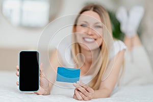 Happy young blond european lady shows smartphone with blank screen and credit card, lies on bed in bedroom