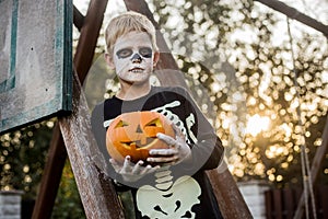 Happy young blond hair boy with skeleton costume holding jack o lantern. Halloween. Trick or treat. Outdoors portrait