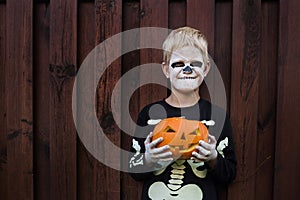 Happy young blond hair boy with skeleton costume holding jack o lantern. Halloween. Trick or treat. Outdoors portrait over wooden