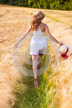 Happy young blonde girl in white dress with straw hat running th