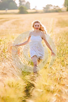 Happy young blonde girl in white dress with straw hat running th