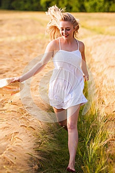 Happy young blonde girl in white dress with straw hat running th