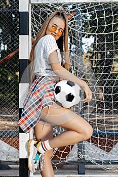 Happy young blonde hipster girl posing with soccer ball in front of goal post at the stadium. Freedom concept.