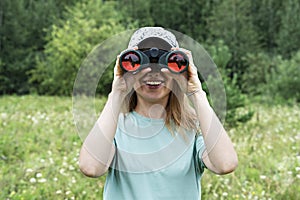 Happy Young blonde woman bird watcher in cap and blue t-shirt looking through binoculars at cloudy sky in summer forest