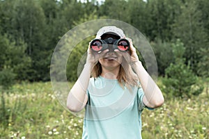 Happy Young blonde woman bird watcher in cap and blue t-shirt looking through binoculars at cloudy sky in summer forest