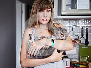Happy young blonde woman in casual t-shirt holding gorgeous scottish fold cat at domestic kitchen. Natural window light portrait.