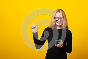 Happy young blonde woman, in glasses, dressed in a strict black dress, holds a smartphone, is typing a text message