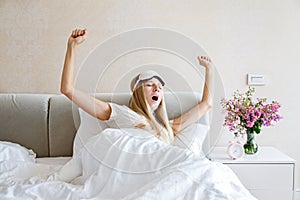 Happy young blonde woman in pajamas and blindfold waking up, yawning and stretching in bed, having a good day. Morning