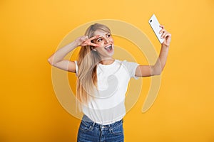 Happy young blonde woman posing isolated over yellow wall background dressed in white casual t-shirt using mobile phone take a