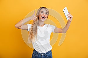 Happy young blonde woman posing isolated over yellow wall background dressed in white casual t-shirt using mobile phone take a